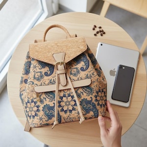 Könnte beinhalten: Ein Rucksack aus Kork mit Klappenverschluss und blauem Blumenmuster. Die Tasche hat einen oberen Griff, verstellbare Träger und eine beige Schnalle. Ein Tablet und ein Smartphone liegen auf dem Tisch.