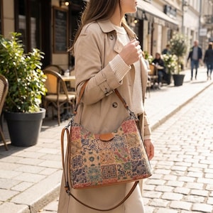 Puede incluir: Bolso de hombro con diseño de patchwork en varios colores y patrones. Tiene una correa de cuero marrón y un cierre de solapa. El bolso se lleva al hombro.