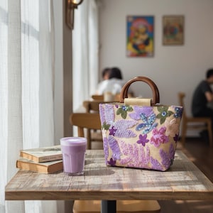 以下が含まれることがあります： 紫、ピンク、緑の色合いの花柄刺繍が施された、木製のハンドルが付いたベージュ色のトートバッグ。 バッグは、紫色の液体が入ったグラスと2冊の本の隣の木製のテーブルの上に置かれています。