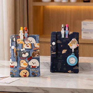 May include: Two rectangular fabric pouches with straps and snap buttons. The left pouch features a blue denim-style print with various dog illustrations and stars. The right pouch is dark blue with a gold line-art pattern, a nurse-themed clip, and a blue analogue watch.