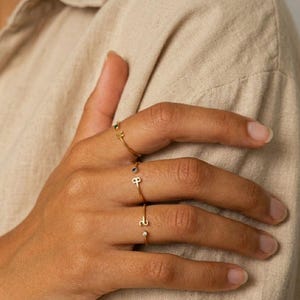 Op de afbeelding: Drie goudkleurige open ringen gedragen aan de vingers. De bovenste ring heeft een kleine letter M, de middelste ring toont een letter P met een minuscuul donker steentje en de onderste ring heeft een letter S met een klein helder steentje.