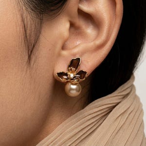 Op de afbeelding: Goudkleurige bloemvormige oorstekers met donkerbruine bloemblaadjes en een klein diamantaccent in het midden. Onder de bloem hangt een grote, ronde, crèmekleurige parel. De oorbellen worden op een oor gedragen.