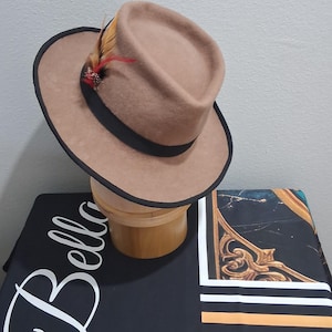 Peut inclure: Un chapeau fedora beige avec un ruban et une bordure noirs, orné de plumes colorées. Le chapeau est présenté sur un support à chapeau en bois, avec une surface à motifs noirs et blancs en arrière-plan.