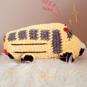 Peut inclure: Un jouet bus scolaire crocheté jaune et gris avec des roues noires et des feux arrière rouges. Le texte BEEP BEEP est écrit en rouge au-dessus du bus. Une ligne jaune avec des flèches indique une longueur d'environ 20 cm pour ce véhicule.