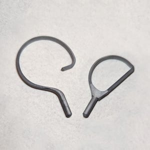 May include: Two gray metal tools are displayed against a white background. One tool is a curved hook shape, and the other is a paddle-like shape with a handle. Both tools have a matte finish and appear to be made of a solid metal.