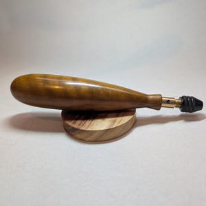May include: A hand drill with a wooden handle and a metal chuck. The handle is a warm brown color and has a smooth, polished finish. The drill is resting on a small, round wooden base.