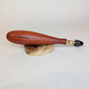 Peut inclure: Un outil manuel doté d'un manche en bois brun rougeâtre lisse avec le texte "JAPANESE TOOLS" imprimé sur le côté. L'outil possède une virole en métal couleur laiton et un petit mandrin cylindrique noir à l'extrémité, posé sur une base en bois.