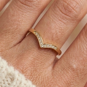 May include: A gold V-shaped ring, set with small, clear gemstones along the top curve. The ring is on a finger, against a cream-coloured knitted fabric. The ring is from the brand "ELARIS JEWELS".