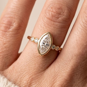 May include: A gold ring featuring a marquise-cut diamond center stone, accented by smaller round diamonds. The ring is set in a gold band. The image also shows the logo of "ELARIS JEWELS WHERE ELEGANCE SHINES".