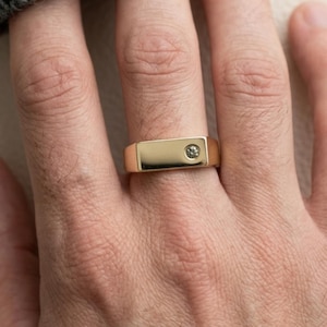 Puede incluir: Un anillo de sello de oro con una superficie superior plana y rectangular que presenta una pequeña piedra preciosa redonda y clara engastada descentrada. El anillo tiene un acabado metálico pulido y liso con un diseño de banda ancha.