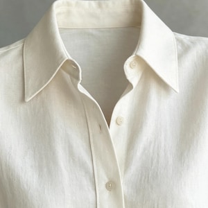 May include: Close-up of a cream-colored button-down shirt. The shirt features a classic collar and a buttoned placket. The fabric appears to be linen or a similar lightweight material, with a slightly textured surface. The buttons are small and round.