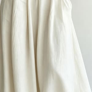 May include: Close-up of a cream-colored fabric with soft folds and a flowing drape. The fabric appears to be a lightweight material, possibly linen or cotton, with a subtle texture. The image highlights the fabric's natural color and the way it catches the light.