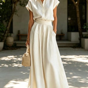May include: A cream-colored linen outfit consisting of a short-sleeved button-down shirt and a flowing maxi skirt. The outfit includes a matching belt and a woven handbag. The ensemble is completed with sandals and sunglasses.