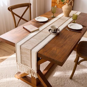 Op de afbeelding: Een houten eettafelset met een beige en witte tafelloper, twee witte borden en een theekopje. De tafelloper heeft een gefranjerde rand en een geweven ontwerp. Twee houten stoelen zijn ook zichtbaar.