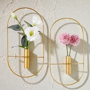Puede incluir: Dos jarrones de pared de metal dorado, cada uno con un marco ovalado y un jarrón cilíndrico. Uno contiene flores blancas y hojas verdes, el otro flores rosas. Los jarrones están montados en una pared clara y texturizada.