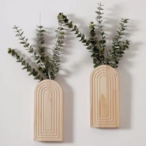 Puede incluir: Dos jarrones de pared de madera clara con diseño arqueado y ranurado, cada uno con ramas de eucalipto verdes. Montados en una pared blanca, crean una estética minimalista y natural.