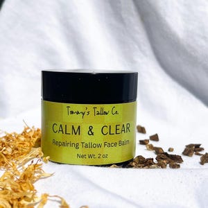 May include: A round jar with a black lid and a yellow label featuring the text Tommy's Tallow Co. CALM & CLEAR Repairing Tallow Face Balm Net Wt. 2 oz. The jar is surrounded by dried yellow flower petals and small dark brown botanical pieces.