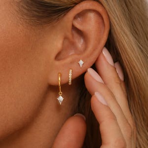 Puede incluir: Primer plano de una oreja adornada con tres pendientes dorados. Un pequeño pendiente de botón, un pequeño aro con un dije en forma de diamante y un pequeño aro con una fila de cristales. Los pendientes están colocados sobre la piel.