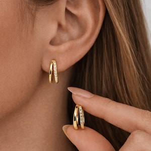 Puede incluir: Pendientes de aro dobles de color dorado. Un aro está adornado con pequeñas piedras brillantes. Los pendientes se muestran en una oreja y son sostenidos por una mano, mostrando su diseño y tamaño. La joya tiene un acabado pulido.