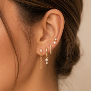 May include: A set of four gold earrings worn in a single ear. The collection includes a stud with a pearl and chain, a small hoop with a pearl and crystal drop, a thin crystal-encrusted hoop, and a small stud featuring a crystal and pearl.