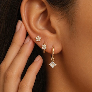 May include: A close-up of three gold earrings with a floral design. One is a stud, another is a small hoop, and the third is a hoop with a dangling flower. Each earring features small, clear gemstones.