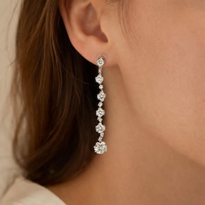 May include: Dangling silver earrings featuring multiple clear, round gemstones. The earrings are attached to a post and cascade downwards in a linear arrangement, with the largest gemstone at the bottom. The earrings are worn on an ear.