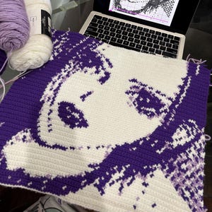 May include: A crocheted portrait in progress, depicting a woman's face in purple and white. The project is on a table with yarn and a laptop showing a pattern. The yarn is in shades of purple and white.