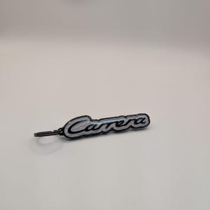 Pode incluir: Um porta-chaves Carrera preto e prateado. A palavra "Carrera" está escrita em itálico com uma contorno prateado. O porta-chaves tem um pequeno anel preto preso no lado esquerdo.