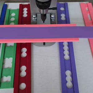 Puede incluir: Una prensa gris, marcada con "1.25 TON", se coloca sobre tiras de espuma artesanal de colores rosa, morado, verde y rojo. Las tiras de espuma están dispuestas sobre una superficie blanca, con varias formas recortadas.