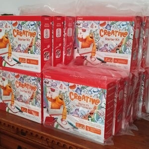 Può includere: Diverse scatole rosse e bianche di Creative Starter Kit sono impilate su una superficie di legno. Le scatole presentano la parola "Creative" in grandi lettere arancioni, insieme a illustrazioni di personaggi dei cartoni animati e strumenti di disegno. Le scatole sono avvolte in plastica trasparente.