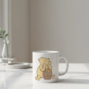 May include: White ceramic mug featuring a cartoon illustration of Winnie the Pooh sitting next to a honey pot. The honey pot is brown with the word "HONNY" printed on it. The mug has a curved handle.