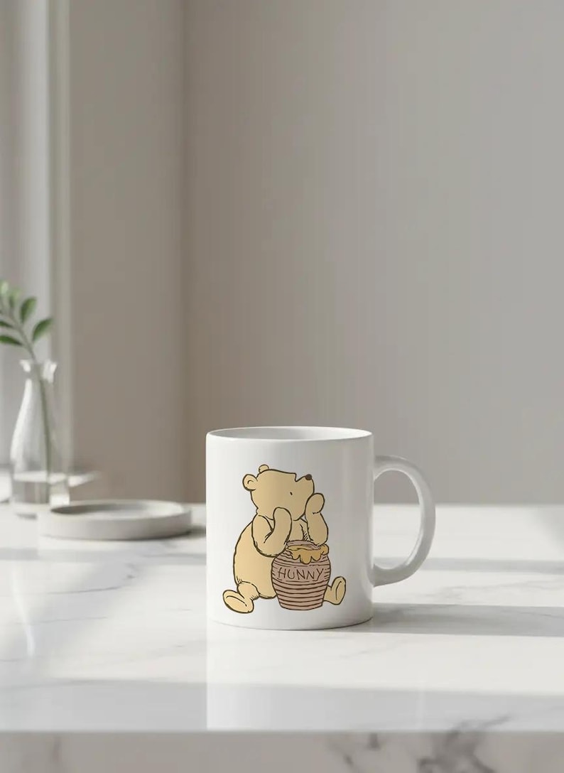 May include: White ceramic mug featuring a cartoon illustration of Winnie the Pooh sitting next to a honey pot. The honey pot is brown with the word "HONNY" printed on it. The mug has a curved handle.