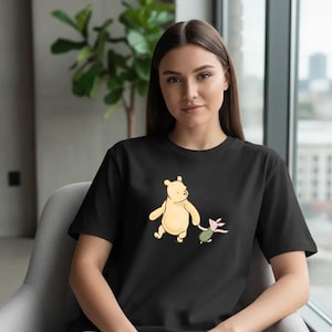 May include: Black t-shirt featuring a cartoon illustration of a yellow bear and a pink pig. The bear is holding the pig's hand. The shirt is a crew neck style.