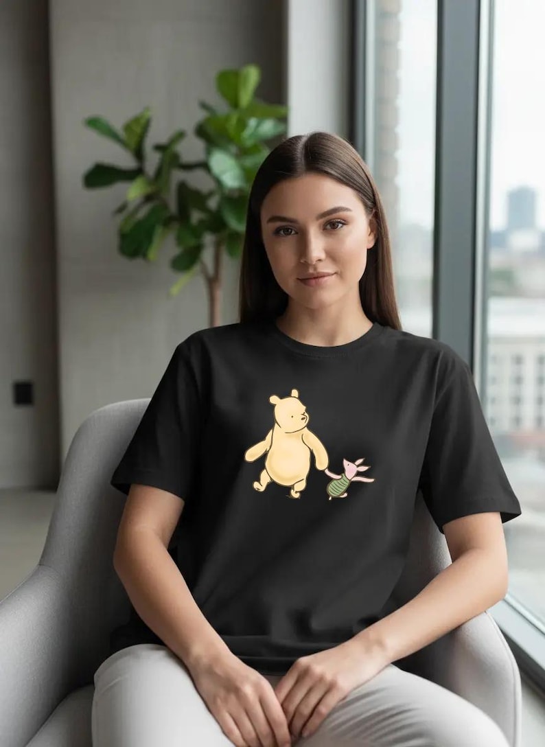 May include: Black t-shirt featuring a cartoon illustration of a yellow bear and a pink pig. The bear is holding the pig's hand. The shirt is a crew neck style.