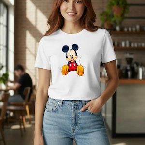 Puede incluir: Camiseta blanca con un dise&ntilde;o de Mickey Mouse. El personaje de dibujos animados tiene orejas negras, una camisa roja, zapatos amarillos y pantalones cortos negros. La camiseta se combina con vaqueros azules.