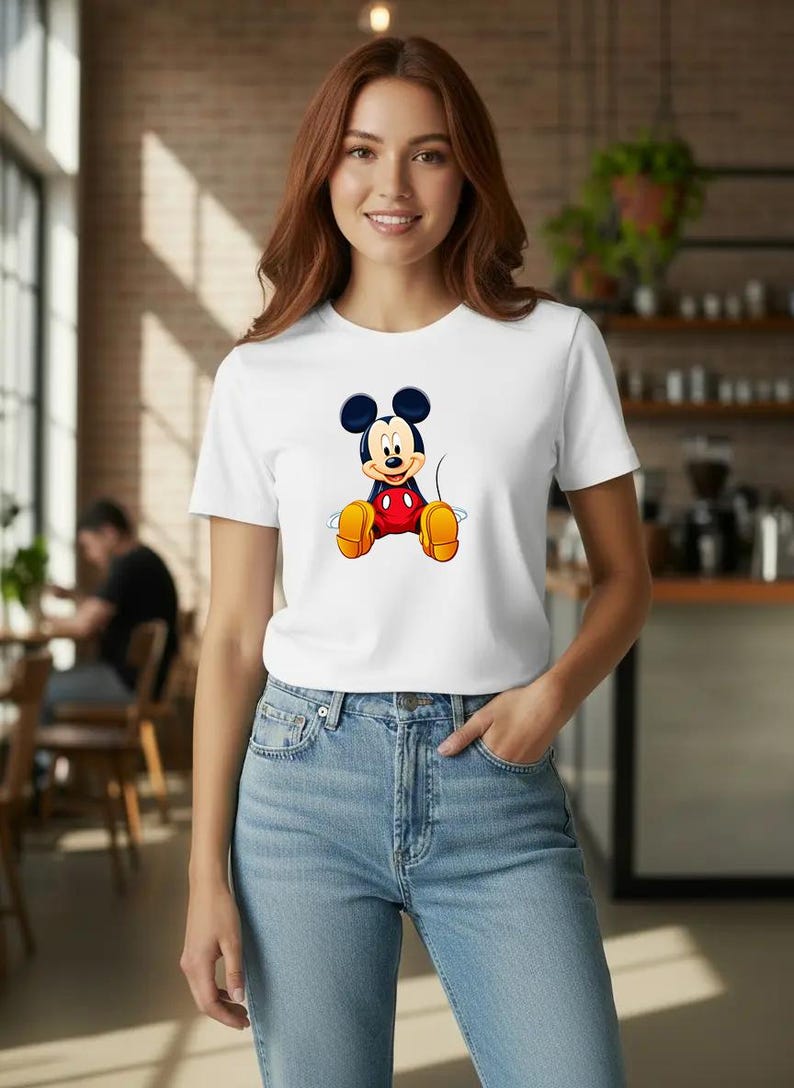 Puede incluir: Camiseta blanca con un dise&ntilde;o de Mickey Mouse. El personaje de dibujos animados tiene orejas negras, una camisa roja, zapatos amarillos y pantalones cortos negros. La camiseta se combina con vaqueros azules.