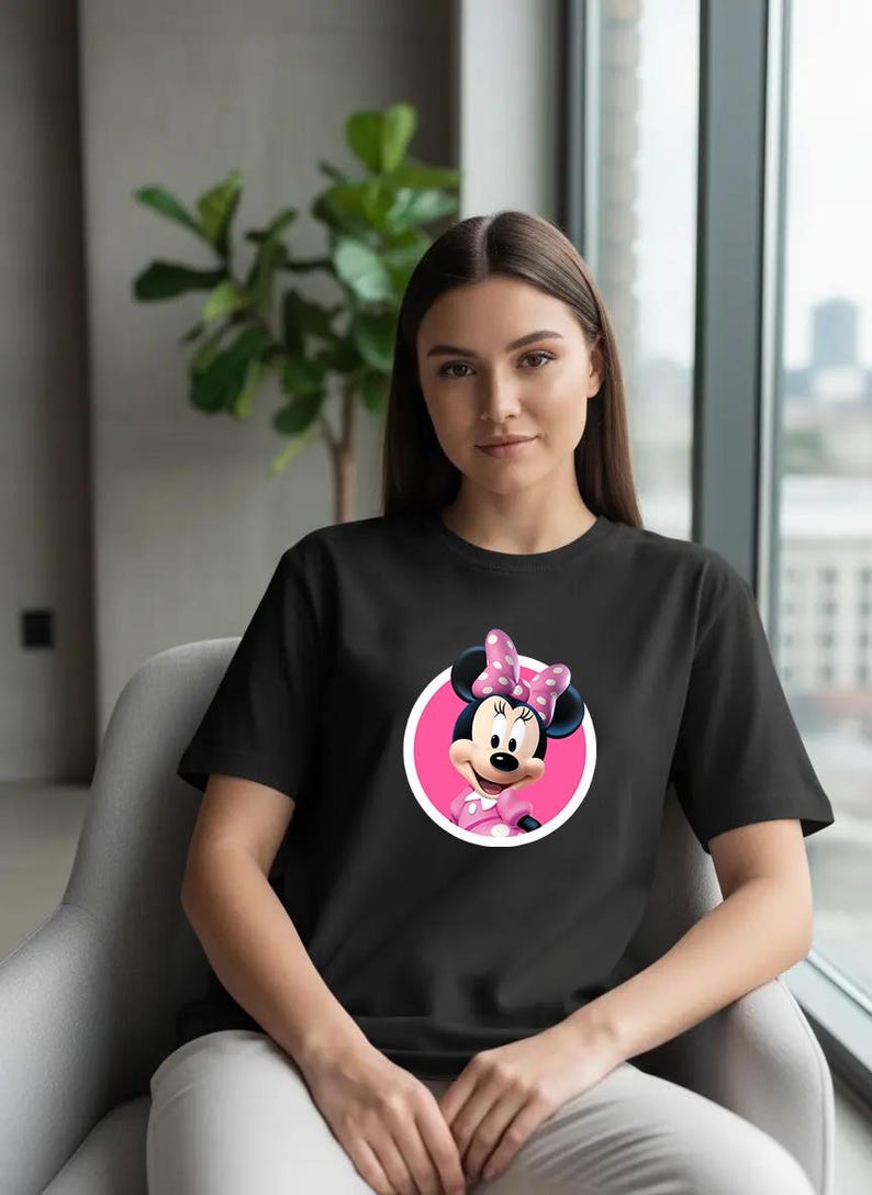 Puede incluir: Camiseta negra con un dise&ntilde;o de Minnie Mouse. El gr&aacute;fico muestra a Minnie Mouse con un lazo y un vestido de lunares rosas, dentro de un c&iacute;rculo rosa. La camiseta la lleva una persona.