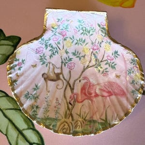 May include: A decorative seashell painted with a tropical scene. The pink shell features a tree with pink and yellow flowers, a sloth, and two pink flamingos. The shell's edge is trimmed with gold.