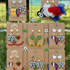 Peut inclure: Une collection de boucles d'oreilles créoles faites à la main sur des supports en carton brun. Les bijoux présentent divers pendentifs dont des ballons de football, des nœuds, des cœurs aux drapeaux nationaux et un sachet de chips Lay's.