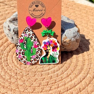 Può includere: Orecchini pendenti con un design cactus e Frida Kahlo. Gli orecchini hanno borchie a forma di cuore rosa, un fiocco verde e un orecchino a forma di goccia con un cactus e stampa leopardata. L'altro orecchino presenta un design Frida Kahlo.