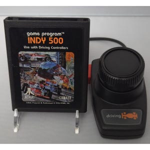 May include: A black Atari Indy 500 game cartridge with a label featuring race cars and the text game program INDY 500 Use with Driving Controllers, positioned next to a black plastic driving controller with a circular knob and a car icon.
