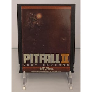 May include: A black rectangular video game cartridge for the Atari 2600 featuring a brown label with the title Pitfall II Lost Caverns in silver and orange text. The label displays an illustration of a character holding a torch in a dark cave.