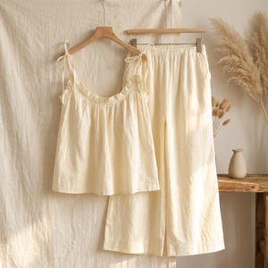 Könnte beinhalten: Cremefarbenes Pyjama-Set. Das Set beinhaltet ein Camisole-Top mit Bindeträgern und einem Rüschenausschnitt sowie eine Hose mit weitem Bein. Die Kleidungsstücke hängen an Holzbügeln vor einem neutralen Hintergrund. Der Stoff scheint leicht zu sein.