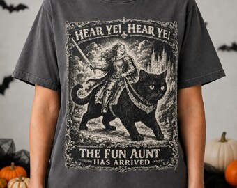 May include: A charcoal gray t-shirt featuring a graphic print of a person in armor riding a large black cat. The design is enclosed in an ornate rectangular frame with the text "HEAR YE! HEAR YE!" at the top and "THE FUN AUNT HAS ARRIVED" at the bottom.