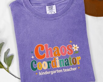 May include: A purple Comfort Colors t-shirt with the words "Chaos Coordinator" in colorful retro font. The text "kindergarten teacher" is below the main text. A daisy and star accents complete the design. The shirt is folded.