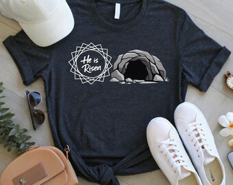 May include: Dark gray t-shirt with the text "He is Risen" and a graphic of an empty tomb. The shirt is paired with white sneakers, a white baseball cap, and a small purse. The design is suitable for Easter.