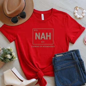 May include: A red t-shirt with the word "NAH" in a square, resembling a periodic table element. The text "ELEMENT OF BOUNDARIES" is below. The shirt is styled with blue jeans, a brown hat, sunglasses, and white shoes.