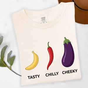 May include: Cream-colored t-shirt featuring illustrations of a banana, chili pepper, and eggplant, with the words "TASTY," "CHILLY," and "CHEEKY" printed below each. The shirt is made of a soft material.