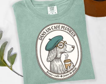 May include: Sage green Comfort Colors t-shirt featuring a circular graphic of a dog wearing a beret and glasses, holding a coffee cup. The graphic includes the text "DANS UN CAFÉ PLUVIEUX" and "livres • café • jours de pluie".
