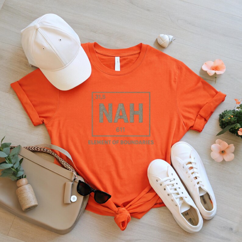 May include: An orange t-shirt with the word "NAH" in a periodic table-style graphic. The shirt is styled with a white baseball cap, white sneakers, a beige purse, and sunglasses. The text "ELEMENT OF BOUNDARIES" is below the graphic.